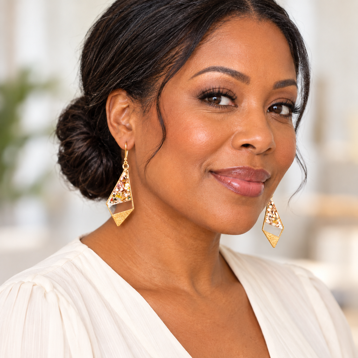 A close-up portrait of a woman wearing gold geometric dangle earrings with colorful floral inlay, paired with a white top against a softly blurred indoor background.
