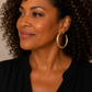 A close-up portrait of a woman with curly brown hair wearing a pair of gold-tone hoop earrings and a black top, set against a plain beige background.