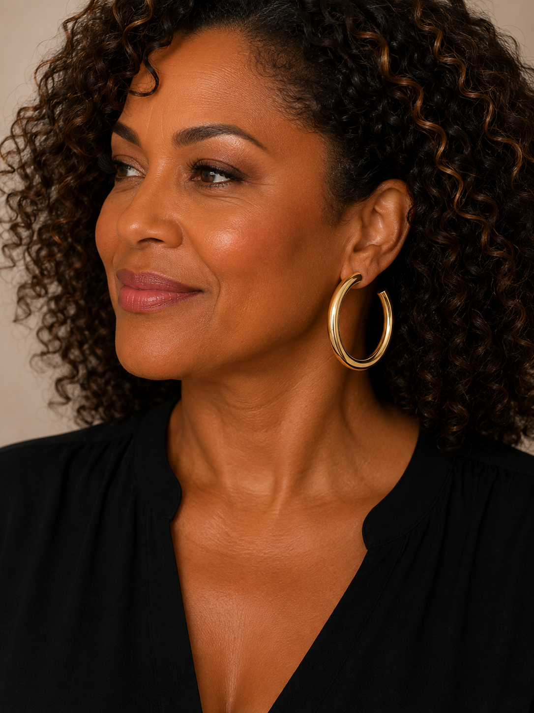 A close-up portrait of a woman with curly brown hair wearing a pair of gold-tone hoop earrings and a black top, set against a plain beige background.