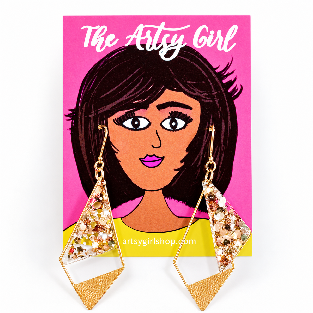 A pair of gold-toned geometric drop earrings with glittery gold and gold triangular charms, displayed on a pink backing card illustrated with a woman's portrait and the text 'The Artsy Girl'.