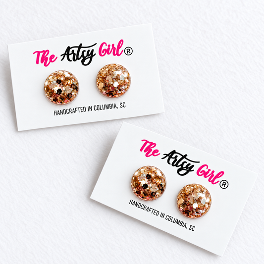 Two round stud earrings covered in rose gold and gold glitter, mounted on white display cards with 'The Artsy Girl' branding, photographed on a white textured background.