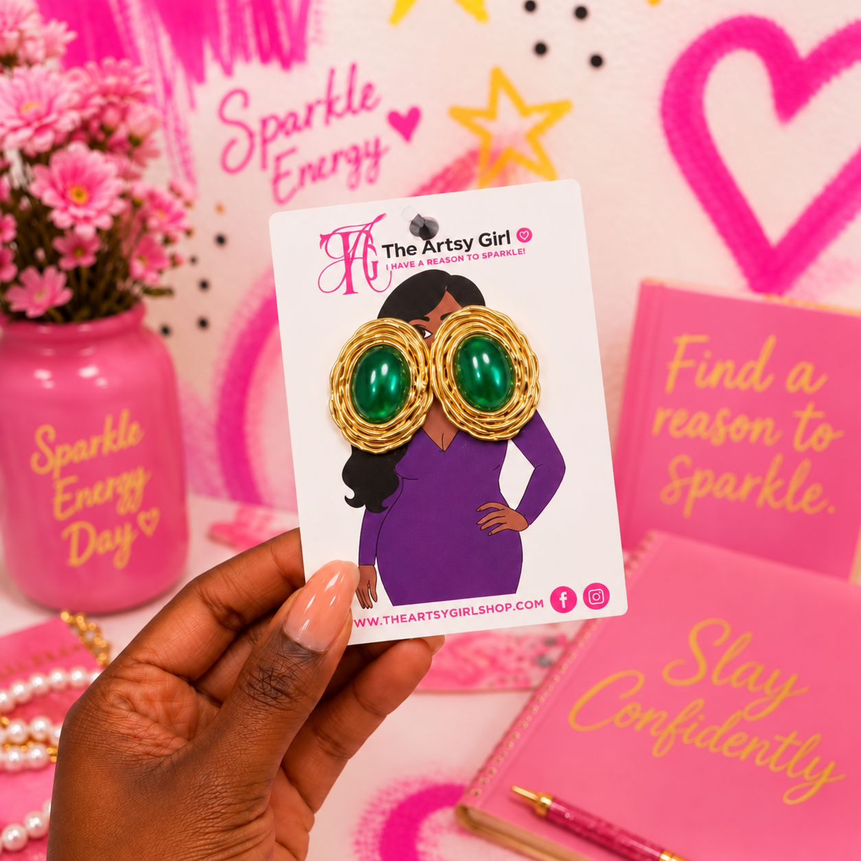 A hand holds a white display card featuring a pair of statement earrings. The earrings have large green oval stones set in gold-toned textured frames.