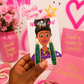 A hand holds a card featuring a hand drawn graphic of a woman wearing green glittery dangle earrings with gold borders. The card is labeled 'The Artys Girl' .