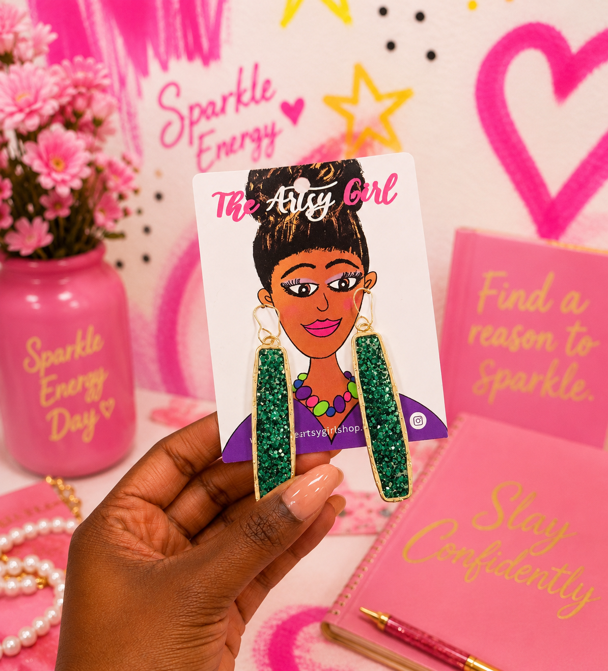 A hand holds a card featuring a hand drawn graphic of a woman wearing green glittery dangle earrings with gold borders. The card is labeled 'The Artys Girl' .
