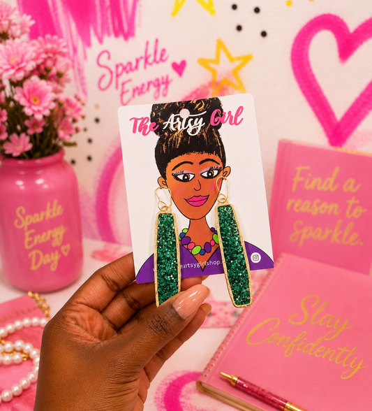 A hand holds a card featuring a hand drawn graphic of a woman wearing green glittery dangle earrings with gold borders. The card is labeled 'The Artys Girl' .