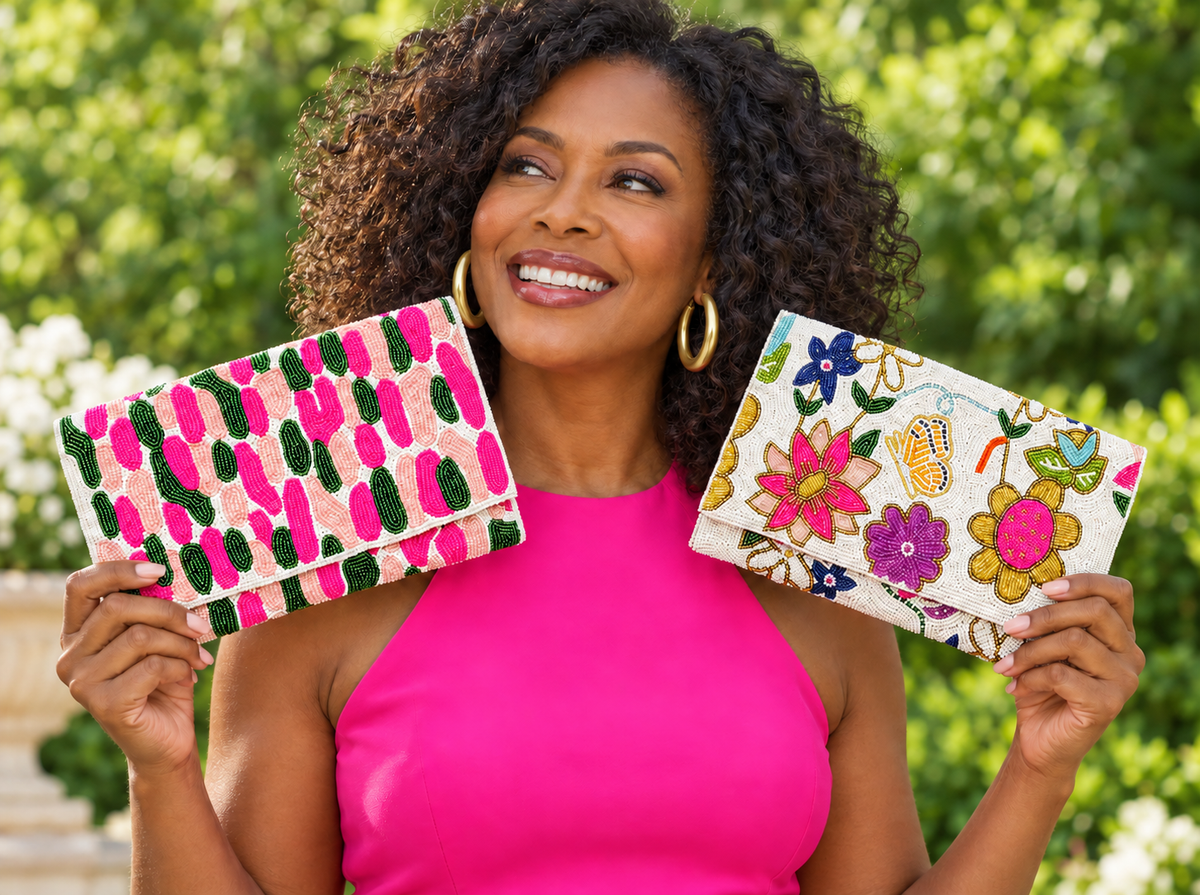 A smiling woman with curly hair holds up two colorful embroidered clutch bags against a blurred green garden background. One clutch has a pink, green, and black abstract pattern, and the other has a white background with pink, yellow, and blue floral embroidery.