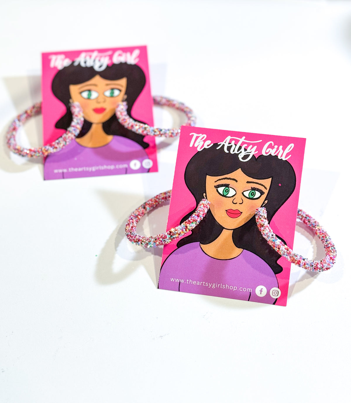 A pair of pink 'Hey Glitter, Hey' hoop earrings with glitter embellishments, displayed on a branded card.