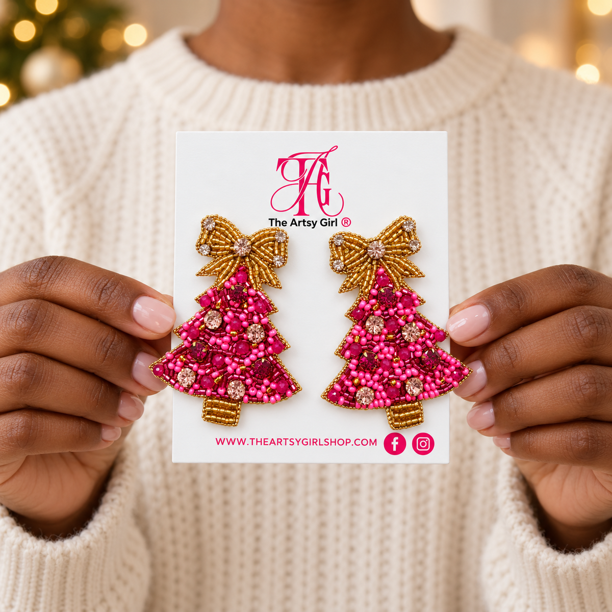 A person wearing a cream knit sweater holds a white card displaying a pair of Christmas tree-shaped beaded earrings. The earrings are made with bright pink and deep red beads, accented with gold-toned beads and rhinestones, and bow details.
