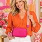 A woman wearing a bright orange blouse and white pants carries a hot pink crossbody bag with a patterned strap and tassel. The bag has an embossed monogram logo on the front.