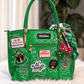 A bright green handbag with multiple decorative patches, gold-tone hardware, a striped green scarf, and an ivy lead bag charm, displayed on a white faux-fur surface with a blurred indoor background.