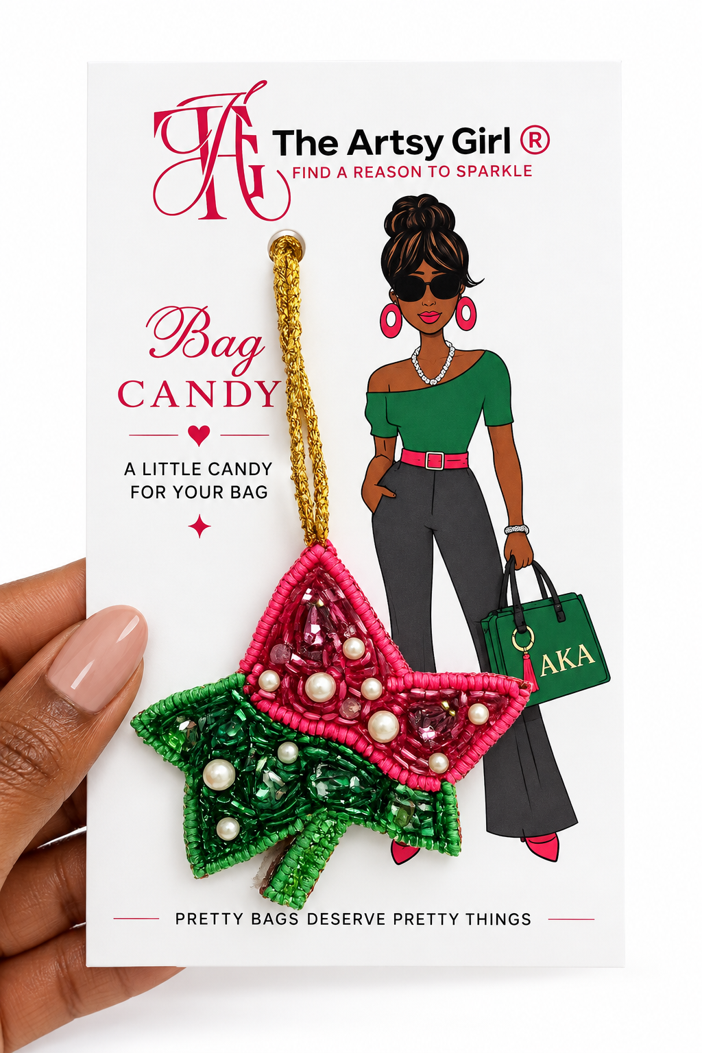 A hand holds a packaged beaded Ivy Leaf bag charm ornament. The ornament is made of green and pink beading with white pearl-like accents and hangs from a gold string.