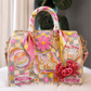 A highly decorated pink and yellow checkered handbag with multiple embroidered patches, a pink satin scarf on the handle, and a red sequined crab embellishment, displayed on a white faux fur surface.