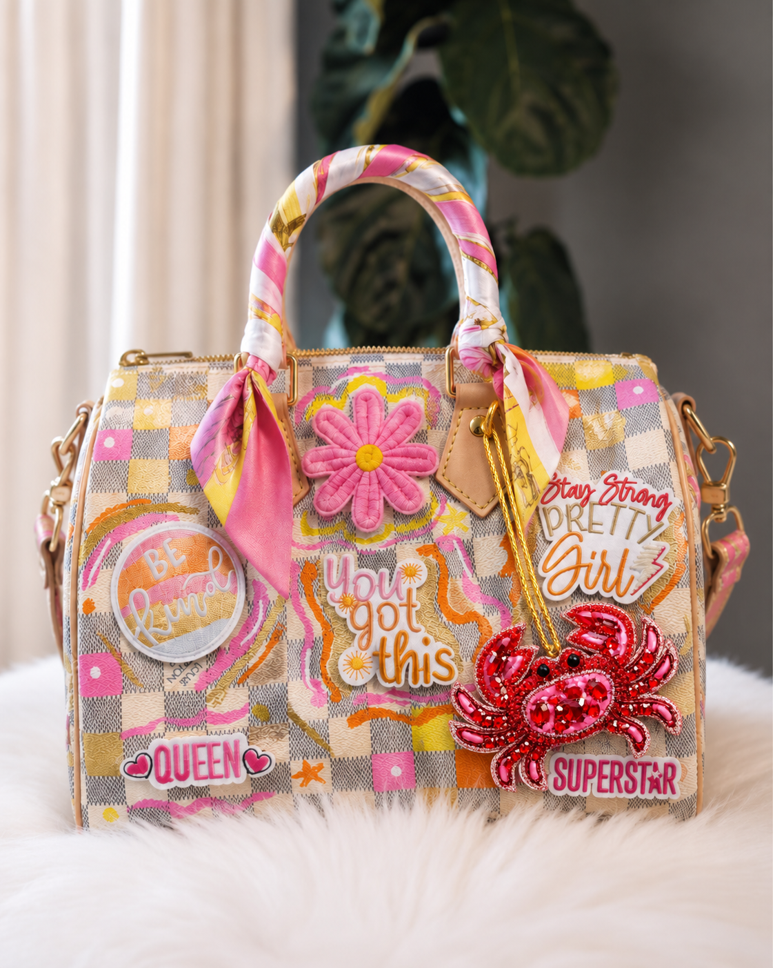 A highly decorated pink and yellow checkered handbag with multiple embroidered patches, a pink satin scarf on the handle, and a red sequined crab embellishment, displayed on a white faux fur surface.