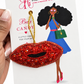 A hand holds a white card featuring a red beaded lip-shaped baby bag charm with a gold hanging loop. The card has branding for 'The Artsy Girl' and includes an illustrated woman in a blue dress holding a green bag.