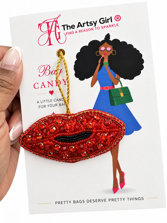 A hand holds a white card featuring a red beaded lip-shaped baby bag charm with a gold hanging loop. The card has branding for 'The Artsy Girl' and includes an illustrated woman in a blue dress holding a green bag.