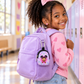 A young girl with curly hair wearing a pink heart-patterned sweater and blue jeans, smiling while looking back over her shoulder. She is wearing a lavender purple backpack with a small round cartoon character accessory hanging from the side pocket, set in a brightly lit school hallway with lockers.
