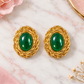 A pair of gold-toned oval earrings with smooth green cabochon centers, displayed on a white marble surface with soft pink floral props.