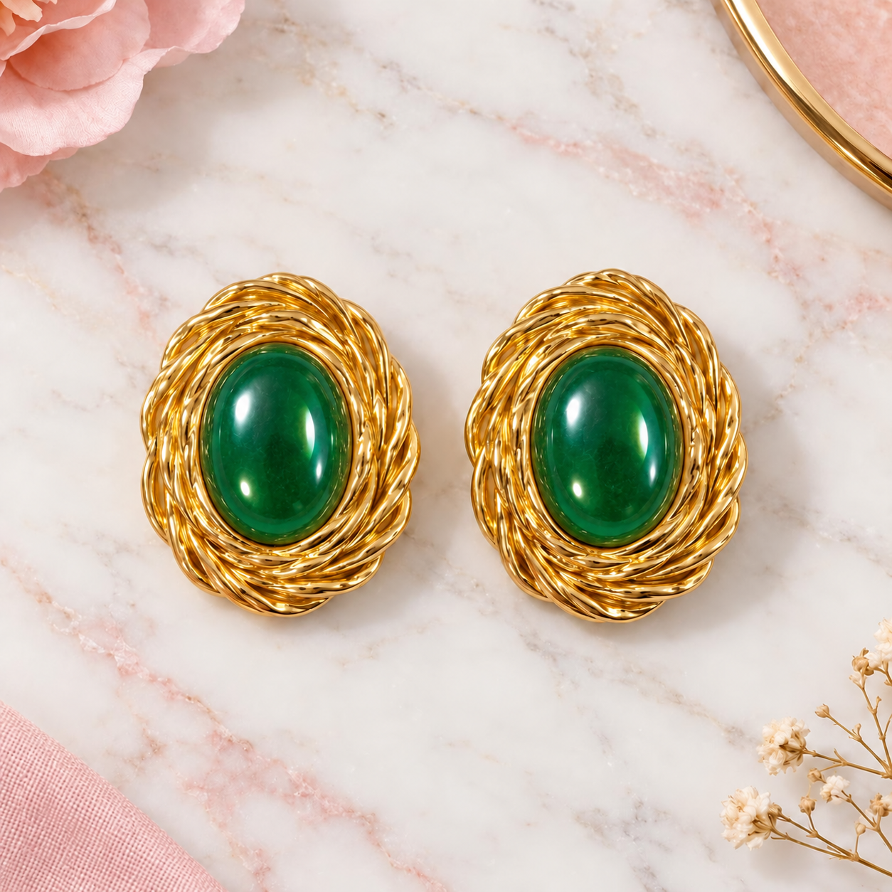A pair of gold-toned oval earrings with smooth green cabochon centers, displayed on a white marble surface with soft pink floral props.