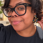 A close-up portrait of a woman wearing Louis Vuitton black cat-eye eyeglasses with small gold star details on the temples, along with colorful beaded earrings. She is wearing a dark navy dress in an indoor setting.
