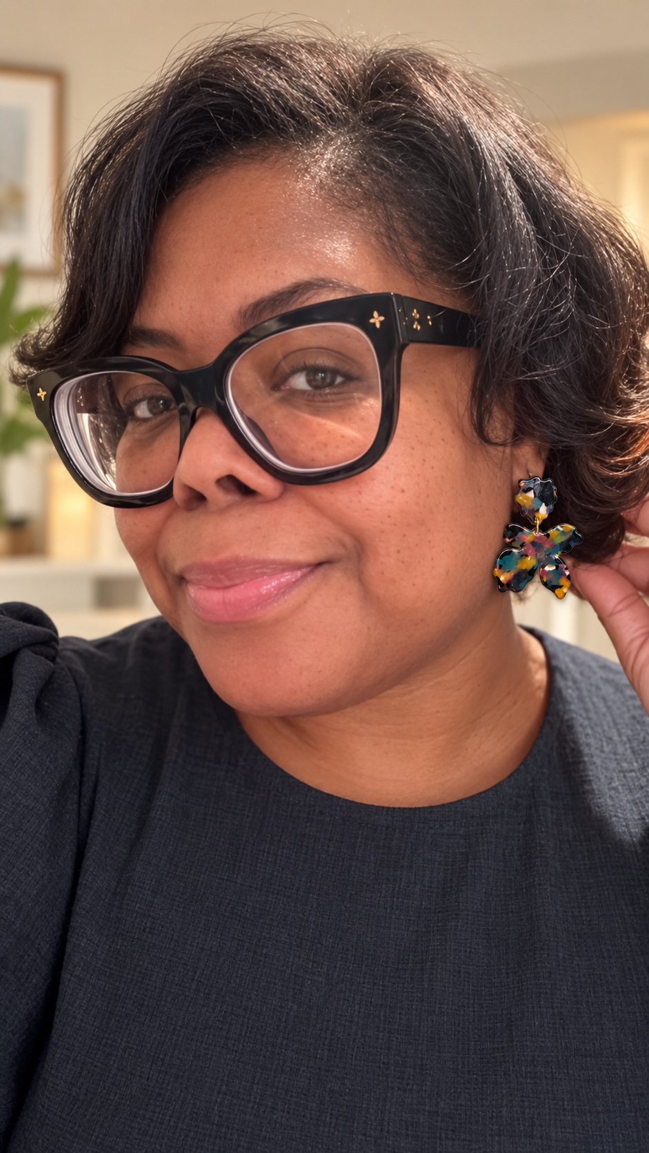 A close-up portrait of a woman wearing Louis Vuitton black cat-eye eyeglasses with small gold star details on the temples, along with colorful beaded earrings. She is wearing a dark navy dress in an indoor setting.