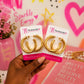 A hand holding two gold hoop earrings on white display cards branded 'The Artsy Girl', against a pink decorative background with hearts, stars, and text.