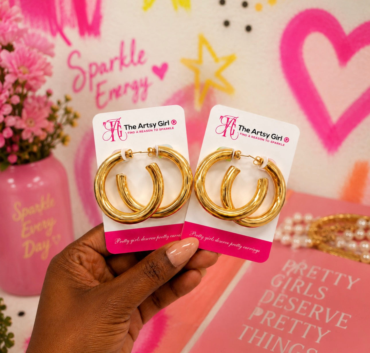 A hand holding two gold hoop earrings on white display cards branded 'The Artsy Girl', against a pink decorative background with hearts, stars, and text.
