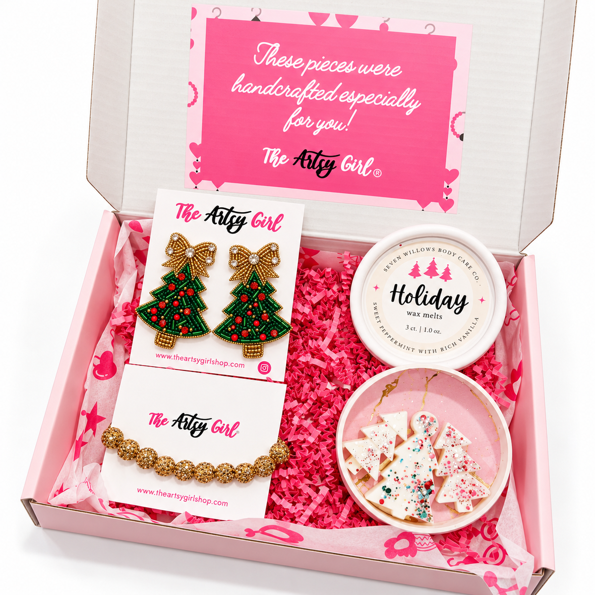 An open pink gift box from The Artsy Girl containing handmade holiday-themed jewelry and candle melts. The box is lined with pink paper and features a pink insert card.