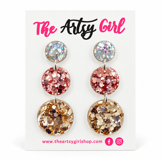 A pair of triple-stone dangle earrings with holographic, pink, and gold glitter discs on silver posts, displayed on a white card labeled 'The Artsy Girl'.