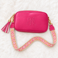 A bright pink crossbody bag with a monogrammed 'A' on the front, featuring a pink and orange patterned woven strap, gold-tone hardware, and a pink and white tassel charm, laid flat on a white faux fur background.