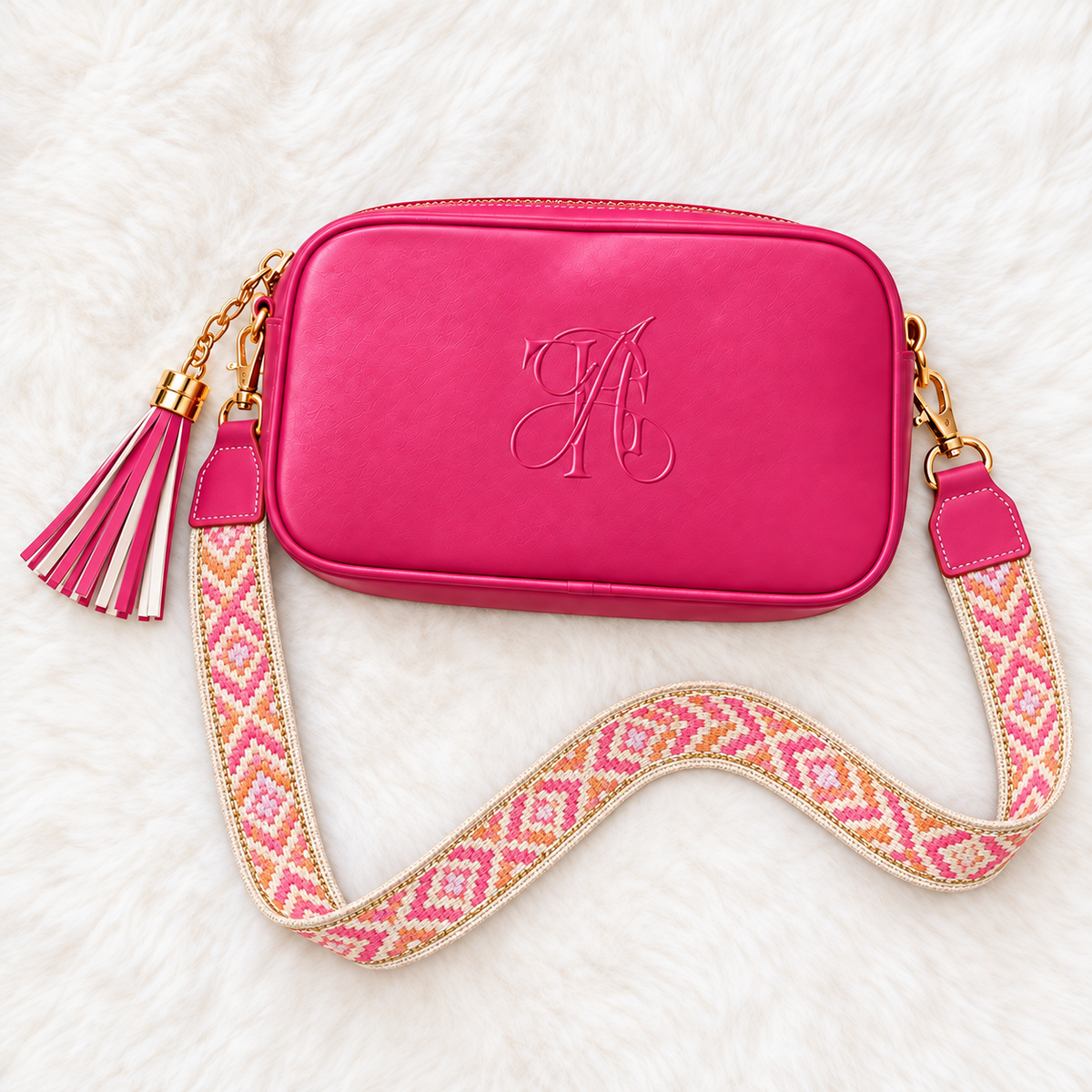 A bright pink crossbody bag with a monogrammed 'A' on the front, featuring a pink and orange patterned woven strap, gold-tone hardware, and a pink and white tassel charm, laid flat on a white faux fur background.