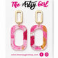 Pink and gold earrings on a card with 'The Artsy Girl' branding.