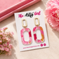 Pink and gold earrings on a 'The Artsy Girl' branded card with pink flowers around.