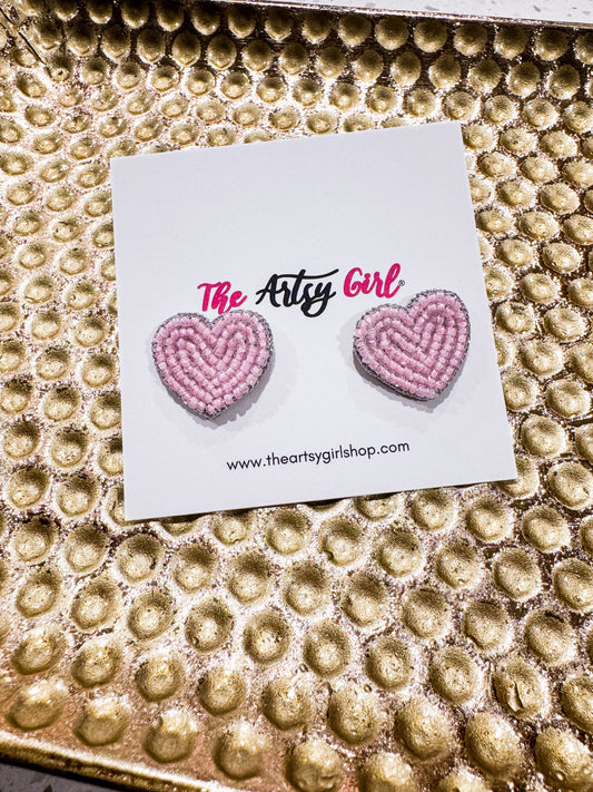 Heart-shaped pink earrings on a card with 'The Artsy Girl' branding on a textured gold background.