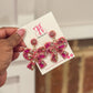 A pair of pink and gold bow-shaped earrings with crystal-clear stones, displayed on a card.