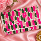 A folded clutch bag with a cactus pattern, featuring bright pink, dark green, and light pink cactus shapes on a white base. The clutch is styled on a pink satin background with decorative pink flowers, a pearl necklace, and a gold flower-shaped dish.