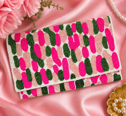 A folded clutch bag with a cactus pattern, featuring bright pink, dark green, and light pink cactus shapes on a white base. The clutch is styled on a pink satin background with decorative pink flowers, a pearl necklace, and a gold flower-shaped dish.