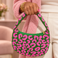 A person wearing a chunky pink knit sweater holds a small crescent-shaped shoulder bag with a bright pink leopard print pattern on a green background. The person is also wearing white pants and gold bracelets.