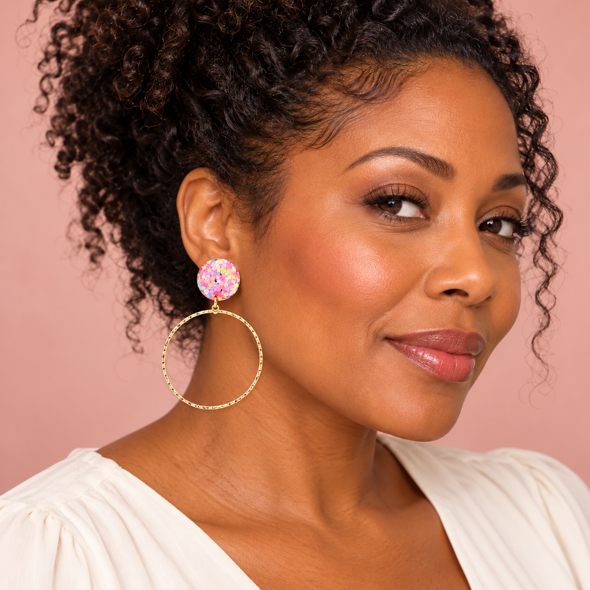 Woman wearing pink pastel confetti glitter studs with dangling gold-tone hoops against a blush background.