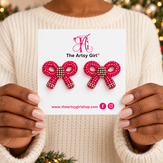 A person in a cream knit sweater holds a white card displaying two pink beaded bow stud earrings with gold rhinestone accents. The card shows the brand name 'The Artsy Girl' and holiday-themed blurred lights in the background.