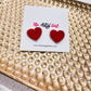 Red heart-shaped earrings on a card with 'The Artsy Girl' branding, placed on a textured gold surface.