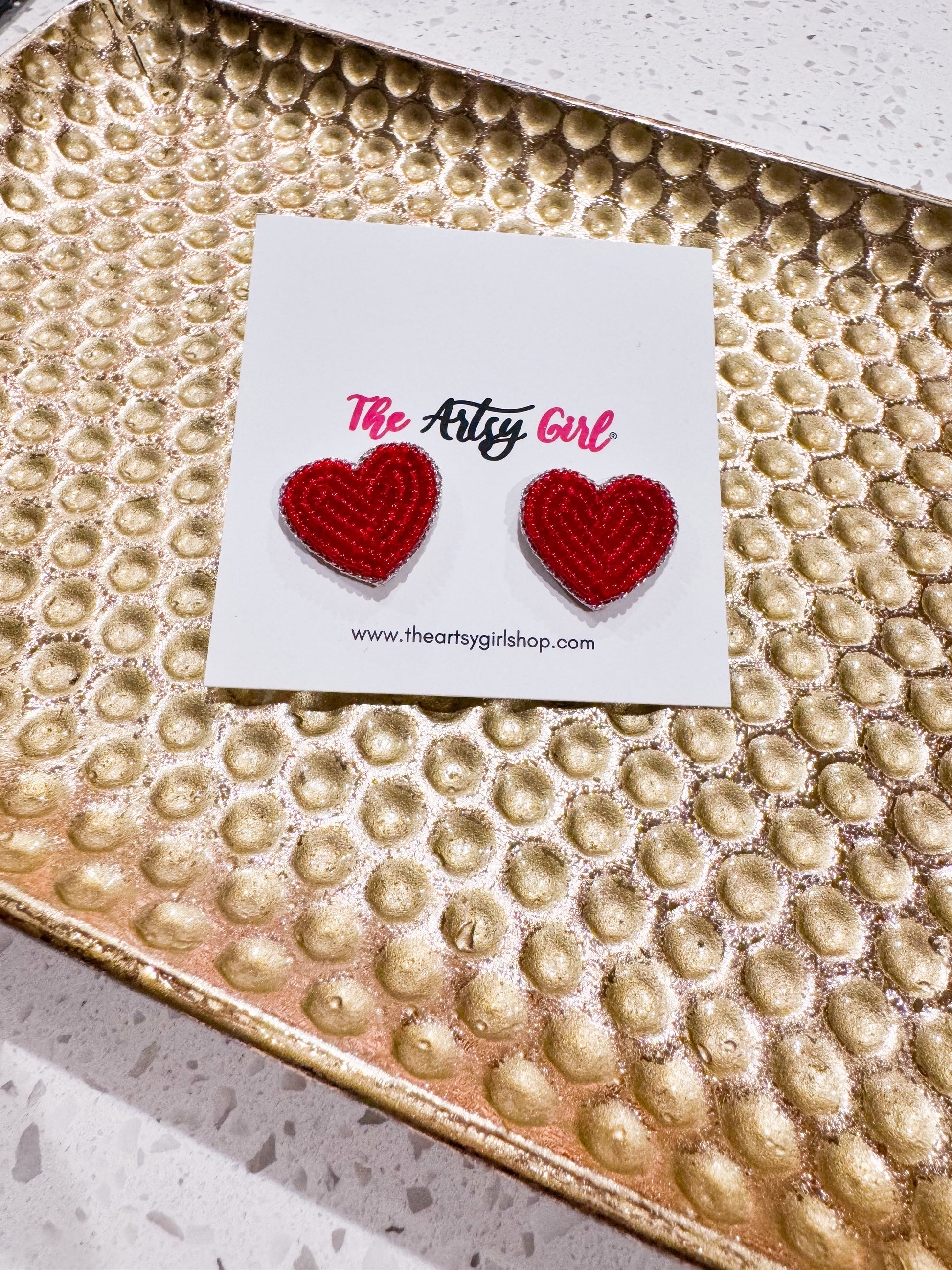 Red heart-shaped earrings on a card with 'The Artsy Girl' branding, placed on a textured gold surface.