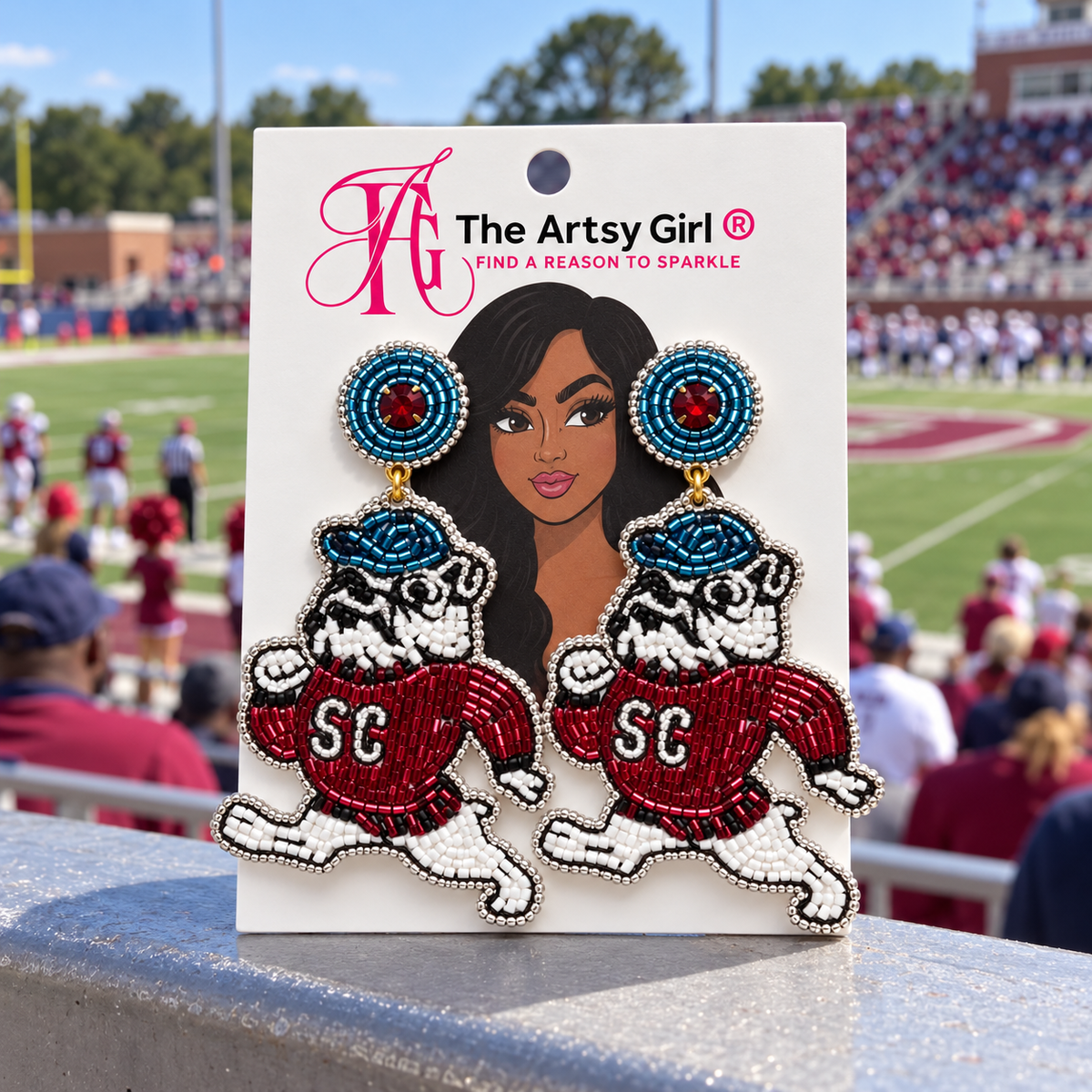 A pair of beaded SC Bulldogs drop earrings on a white display card from 'The Artsy Girl', set against a blurred background of a football stadium.