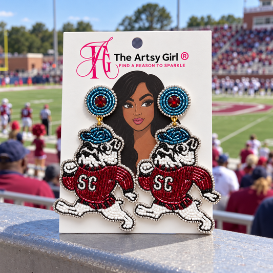A pair of beaded SC Bulldogs drop earrings on a white display card from 'The Artsy Girl', set against a blurred background of a football stadium.