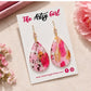 Pink and gold teardrop earrings on a 'The Artsy Girl' branded card with flowers around.