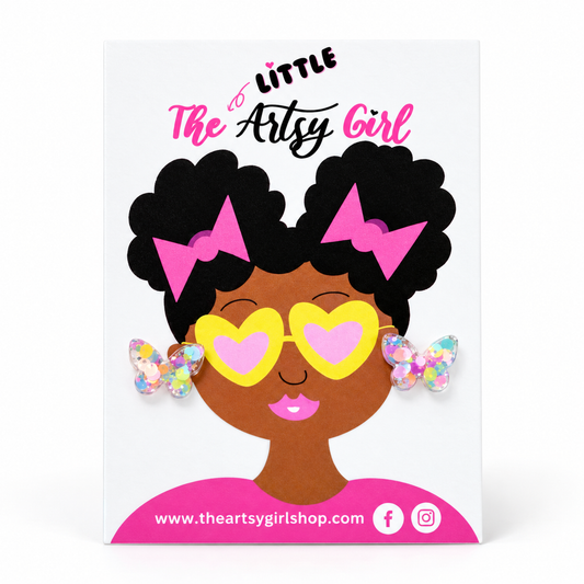 A white display card featuring a cartoon illustration of a Black girl with afro hair, pink bows, yellow heart-shaped sunglasses, and butterfly earrings. The card reads 'Little The Artsy Girl' and includes the website. 