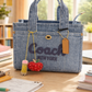 Denim handbag with 'Coach New York' branding on a desk with The Artay Girl Teacher Bag Charm