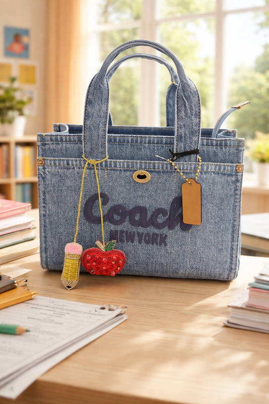 Denim handbag with 'Coach New York' branding on a desk with The Artay Girl Teacher Bag Charm