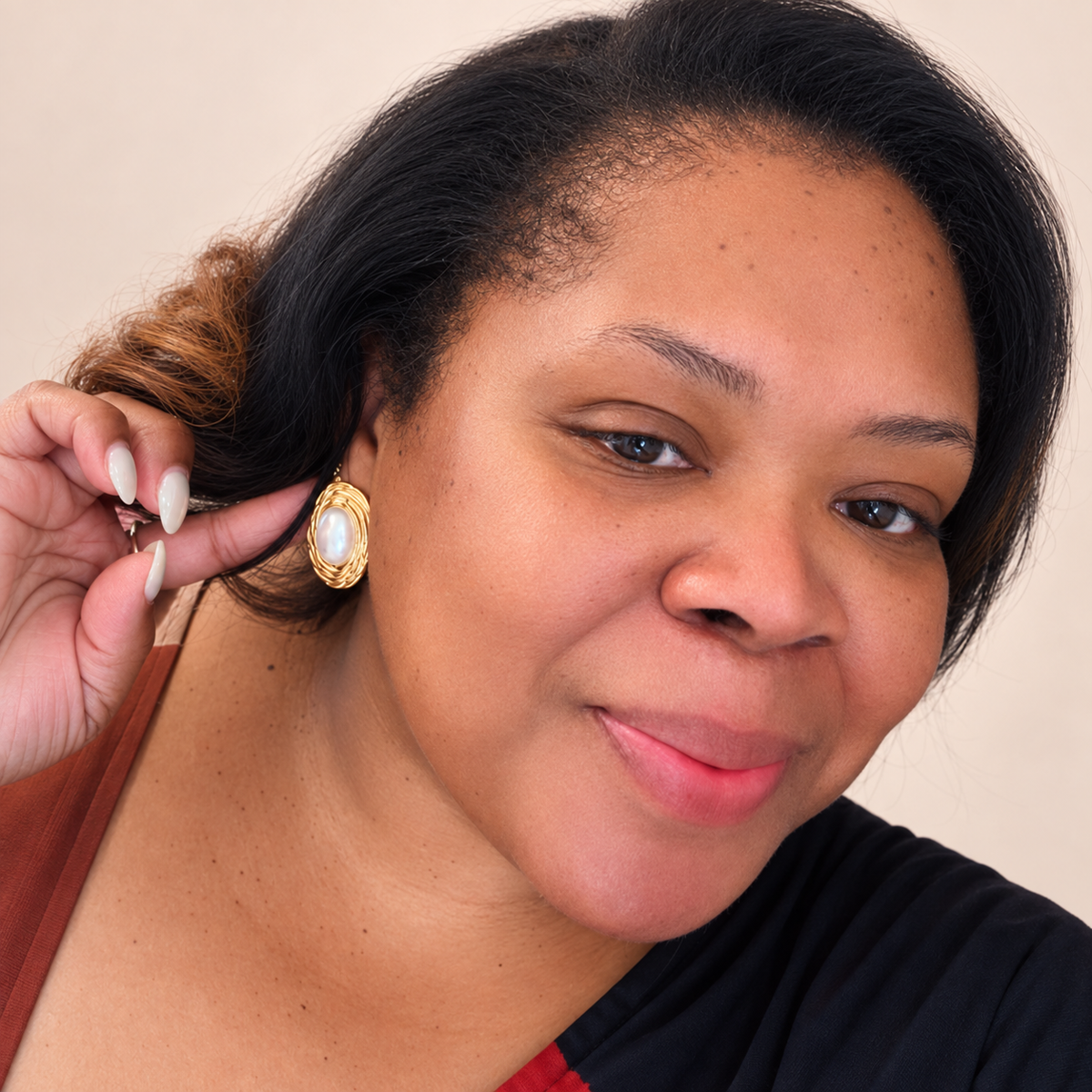A close-up portrait of a woman wearing a gold oval earring with a white pearl center, holding her hair to show the earring more clearly against a plain beige background.