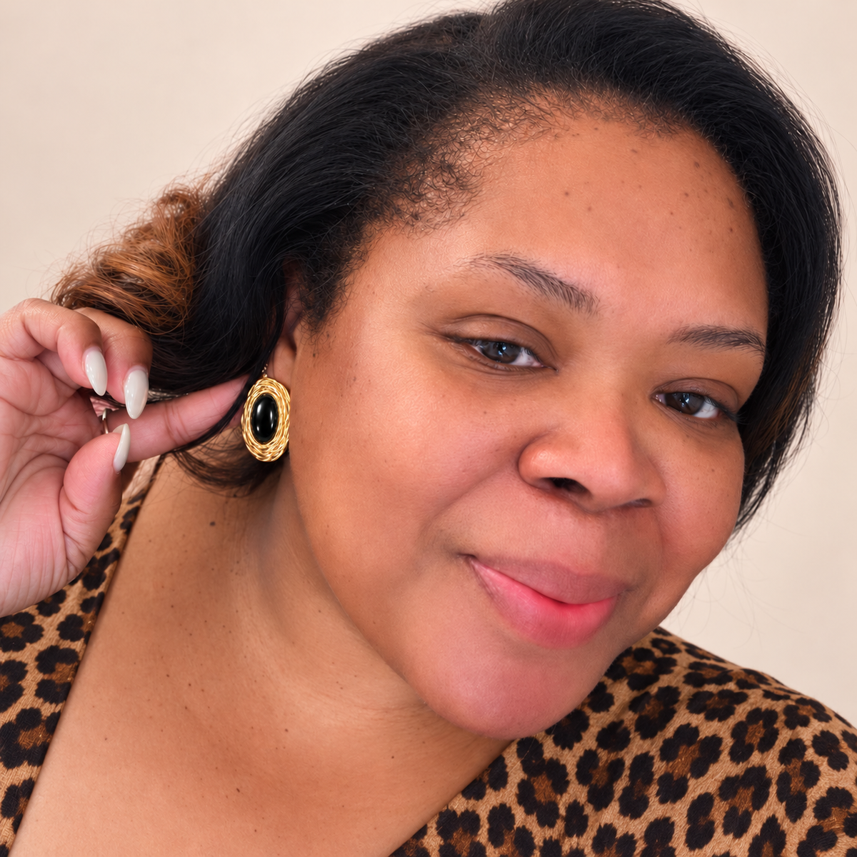 A close-up portrait of a woman wearing a gold-toned oval earring with a black center, holding her hand near her ear. She is wearing a leopard-print top against a plain beige background.