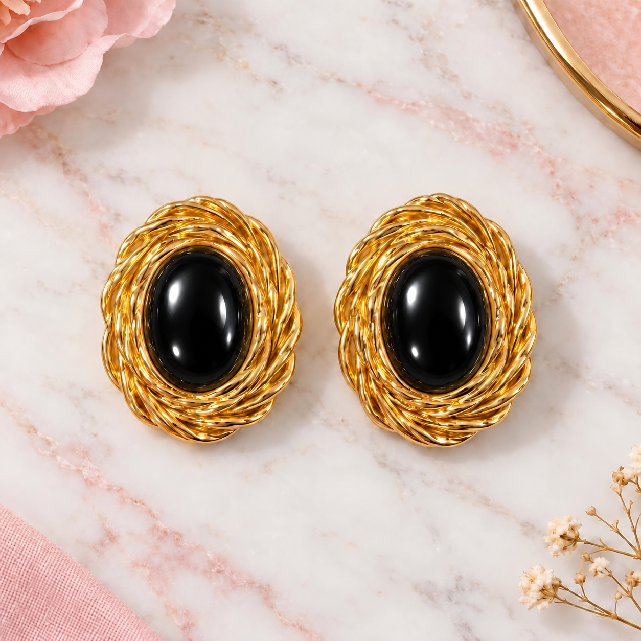 A pair of gold-toned twisted wire earrings with oval black onyx centers, arranged on a white marble surface with soft pink floral props.